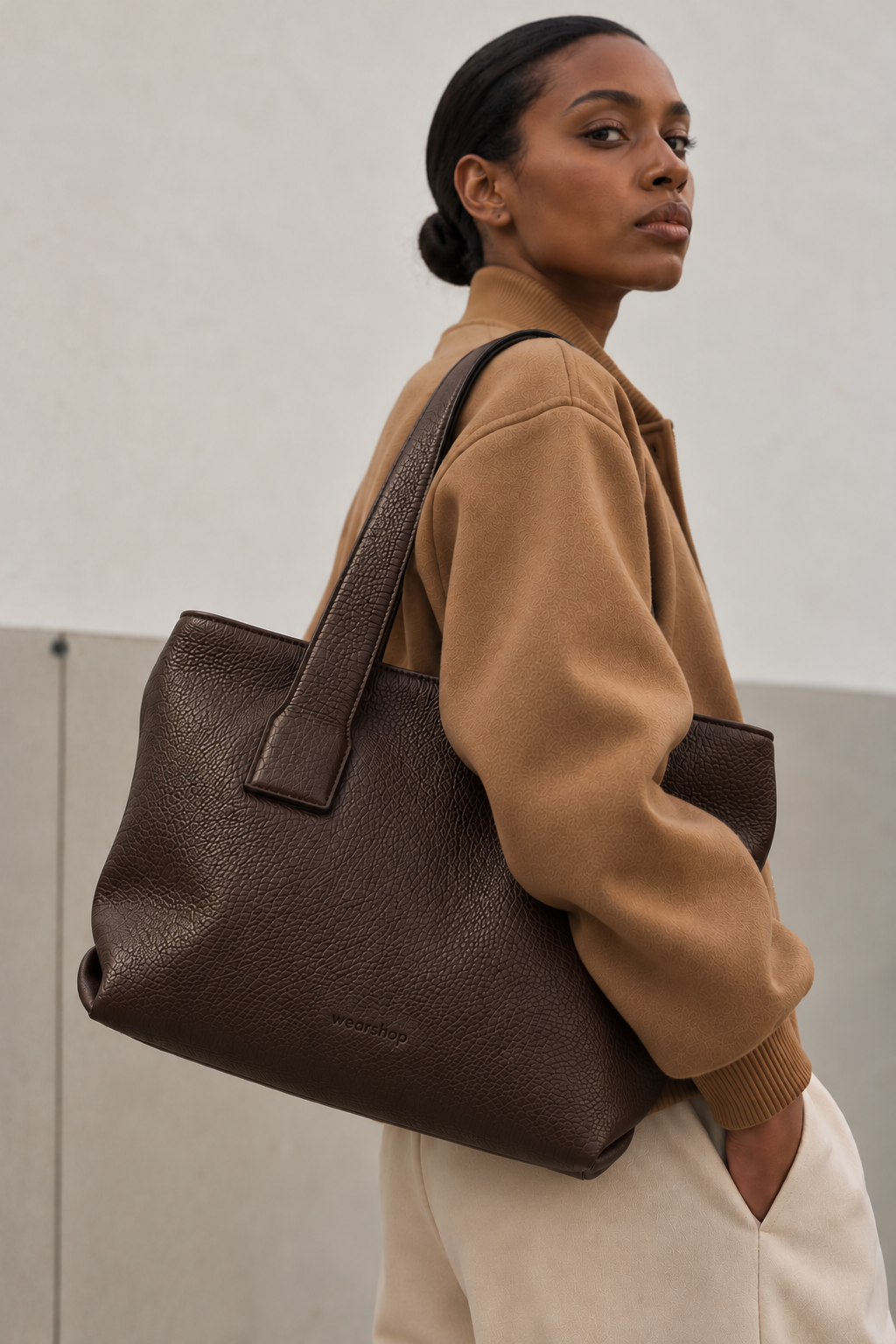 A woman wearing a camel jacket and cream pants carries a large dark brown pebbled leather tote bag over her shoulder, set against a minimalist light gray concrete wall.